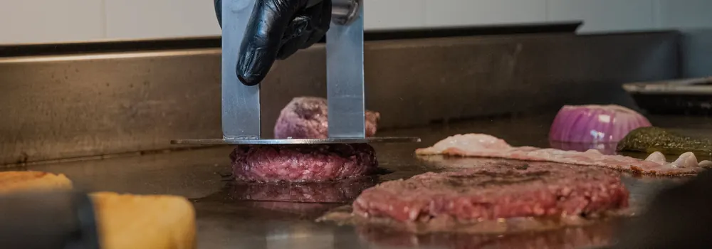 Mustas kindas käsi surub metallpressiga kotletit kuumal grillplaadil