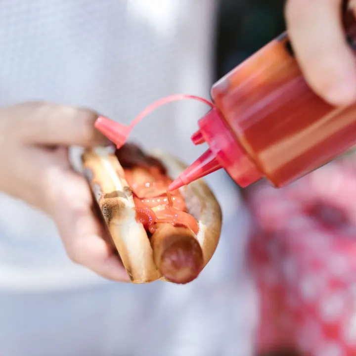 Punane kastmepudel lisab ketšupit käes hoitud hotdogi peale