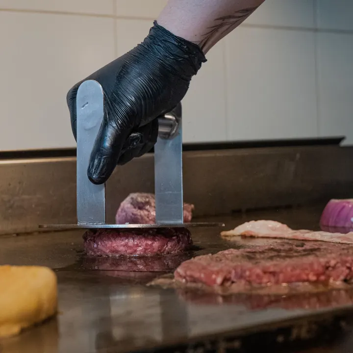 Mustas kindas käsi surub kotletit metallpressiga kuumal grillplaadil